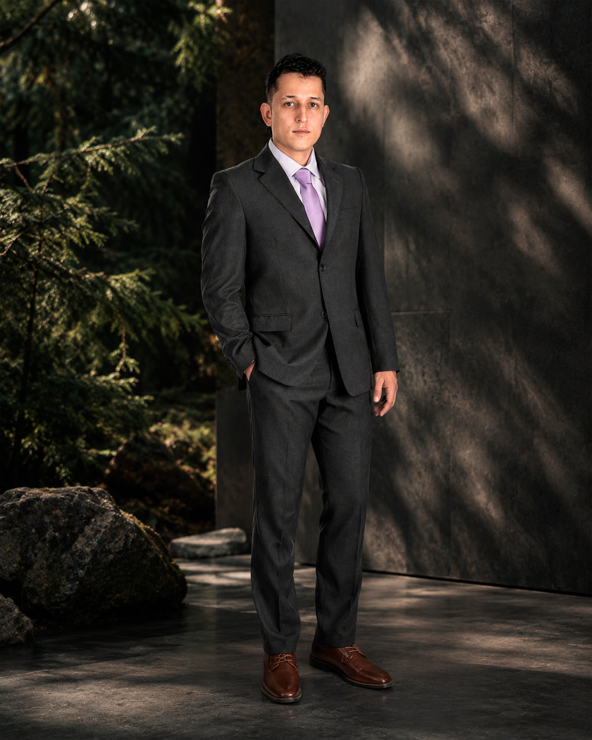 Hombre con traje gris y camisa lila, de pie en un jardín